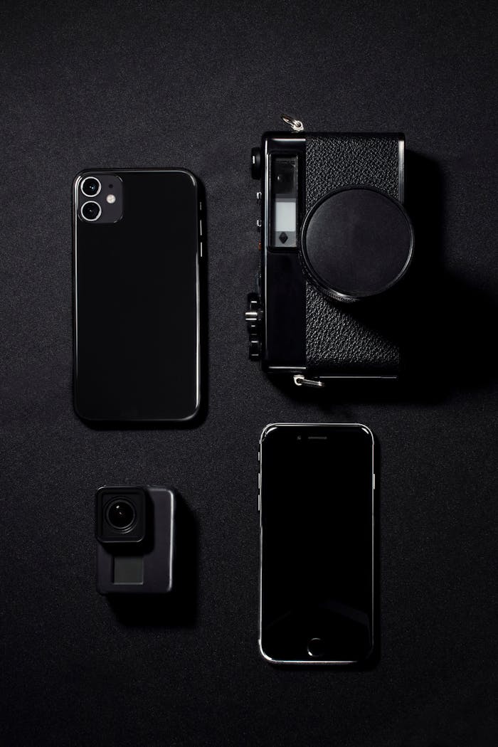 trusted-partner Minimalist flat lay of black gadgets including phones and a camera against a dark background.